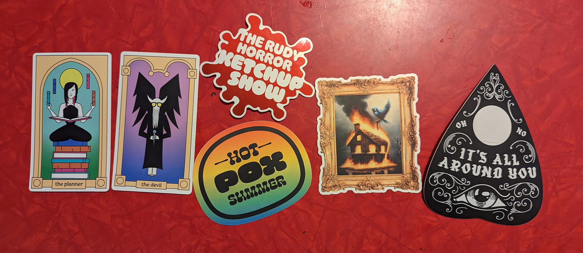 a sample of six stickers from the Says Who Sticker Club including two tarot cards of Maureen and Dan, a splat that reads The Rudy Horror Ketchup Show, one that reads Hot Pox Summer, a framed painting of Twitter burning down, and a ouiji puck that reads It's All Around You